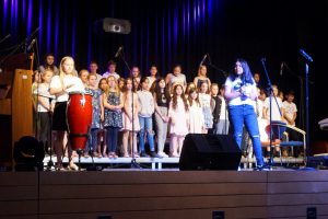 Eine Gruppe von Kindern und Jugendlichen steht auf einer Bühne. Im Vordergrund sind zwei Mädchen zu sehen: eines spielt Congas und das andere spricht ins Mikrofon. Im Hintergrund stehen weitere Kinder in bunten Kleidern, die zusammen singen. Die Bühne ist mit Mikrofonen ausgestattet und von blauer und lila Beleuchtung umgeben.