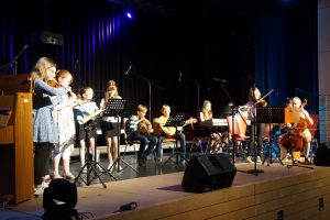 Eine Gruppe von Kindern und Jugendlichen spielt auf einer Bühne verschiedene Musikinstrumente, darunter Flöten, Gitarren, Geigen und ein Cello. Sie sind in einem gut beleuchteten Raum mit einem dunklen Hintergrund und Mikrofonen aufgestellt. Die Musiker sind in verschiedenen Positionen angeordnet und konzentrieren sich auf ihre Darbietung.