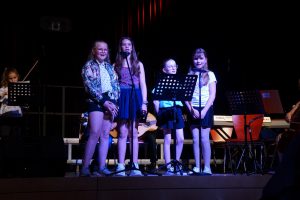 Vier Mädchen stehen auf einer Bühne und singen. Sie halten Mikrofone und schauen in die Kamera. Im Hintergrund sind Musiker zu sehen, die verschiedene Instrumente spielen. Die Bühne ist beleuchtet und die Atmosphäre wirkt festlich.
