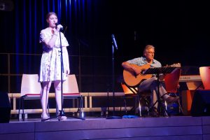 Eine junge Frau steht auf einer Bühne und singt in ein Mikrofon, während ein Mann neben ihr auf einer Gitarre spielt. Die Bühne ist von sanftem Licht beleuchtet und im Hintergrund sind Stühle sichtbar.