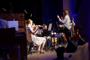 Eine junge Musikerin spielt auf einer Geige, während eine Dirigentin das Orchester leitet. Im Hintergrund sind weitere Musiker mit verschiedenen Instrumenten zu sehen. Die Szene ist auf einer Bühne mit gedämpftem Licht, wobei die Musiker in Formationen angeordnet sind.