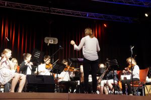 Ein Orchester mit jungen Musikern sitzt auf der Bühne und spielt Instrumente, während eine Dirigentin vor ihnen steht und das Stück leitet. Die Bühne ist schwach beleuchtet, mit bunten Lichtern im Hintergrund. Die Musiker haben Notenständer vor sich und konzentrieren sich auf die Aufführung.
