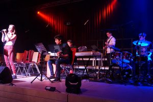 Eine Band tritt auf einer Bühne auf. Eine Sängerin steht vorne und singt ins Mikrofon, während vier junge Musiker sie begleiten: ein Gitarrist, ein Keyboarder und ein Schlagzeuger. Die Bühne ist in warmes Licht getaucht, und Stühle sind im Hintergrund aufgestellt.