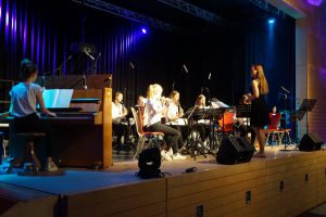 Eine Musikgruppe spielt auf einer Bühne. Im Vordergrund sitzt eine Person am Klavier. Auf der Bühne stehen mehrere Musikerinnen und Musiker mit verschiedenen Instrumenten, darunter Blasinstrumente. Eine Dirigentin leitet die Gruppe. Die Bühne ist beleuchtet, und der Hintergrund zeigt dunkle Vorhänge.