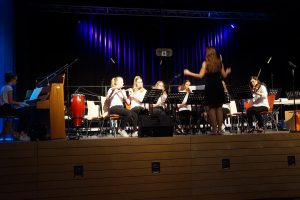 Eine Musikgruppe spielt auf einer Bühne. Im Vordergrund ist eine Dirigentin zu sehen, die mit den Händen dirigiert. Um sie herum sitzen Musikerinnen, die verschiedene Instrumente wie Geigen und Percussion spielen. Im Hintergrund ist eine dunkle Bühne mit blauen Lichtern.