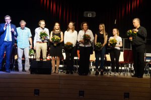 Eine Gruppe von sechs Personen steht auf einer Bühne und hält Blumensträuße in den Händen. Im Hintergrund sind Stühle und eine Bühne mit rotem Vorhang sichtbar. Links spricht eine Person mit Mikrofon, während die anderen in formeller Kleidung dastehen.