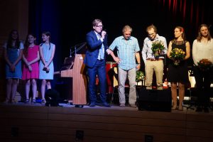 Eine Gruppe von sechs Personen steht auf einer Bühne. Ein Mann in einem Anzug hält ein Mikrofon und spricht. Neben ihm stehen fünf weitere Personen, die unterschiedlich gekleidet sind. Einige von ihnen halten Blumensträuße. Im Hintergrund sind Bühnenbeleuchtung und ein Klavier sichtbar.