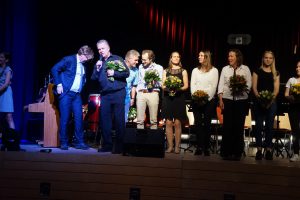 Eine Gruppe von Personen steht auf einer Bühne bei einer Veranstaltung. Einige Personen halten Blumensträuße in den Händen. Im Hintergrund sind Instrumente und eine Bühne mit rotem Licht zu sehen. Die Stimmung wirkt feierlich.