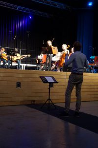 Eine Aufführung in einem Veranstaltungsraum zeigt eine Gruppe von Musikern auf der Bühne, die verschiedene Instrumente spielen. Im Vordergrund steht ein Dirigent, der die Musiker anleitet. Die Bühne ist mit Holzpaneelen umrandet, und die Beleuchtung ist gedämpft, mit blauen Lichtakzenten. Sitzplätze für das Publikum sind im Hintergrund sichtbar.