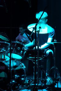Ein junger Mann spielt Schlagzeug auf einer Bühne, während ein weiterer Musiker im Hintergrund zu sehen ist. Die Szene ist schwach beleuchtet, mit einem bläulichen Licht, das den Schlagzeuger hervorhebt. Der Musiker im Hintergrund ist teilweise verdeckt und spielt ein anderes Instrument.