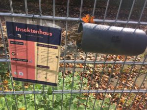 Ein Insektenhaus an einem Zaun. Links hängt ein Informationsschild mit dem Titel 