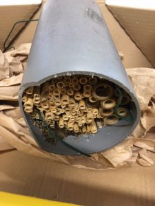 Ein durchgeschnittener grauer Rohrabschnitt, der innen mit vielen kleinen, runden Röhren gefüllt ist. Diese Röhren sind aus einem hellen Material und variieren in der Größe. Der Rohrabschnitt liegt auf einem Haufen von braunem Papier in einer Kartonbox. Es sind einige Drähte sichtbar, die aus dem Rohr herausragen.
