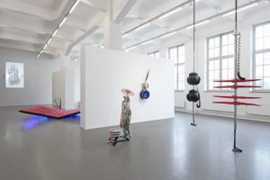 Ein moderner Ausstellungsraum mit weißen Wänden und großen Fenstern. In der Mitte des Raumes stehen verschiedene Kunstwerke, darunter eine Skulptur und mehrere Objekte, die an Wänden und von der Decke hängen. Der Boden ist grau und es gibt eine rote, gestreifte Fläche auf dem Boden, die in den Raum hineinragt. Einige Werke sind mit Lichtakzenten versehen.