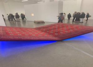 Ein roter Teppich mit traditionellen Mustern ist schräg aufgestellt und hebt sich an einer Ecke, während eine blaue Beleuchtung darunter leuchtet. Im Hintergrund sind mehrere Personen in einem Kunst-Ausstellungsraum zu sehen.