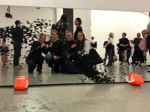 Eine Gruppe von vier Personen posiert vor einem Spiegel in einem Ausstellungsraum. Im Hintergrund sind weitere Besucher zu sehen, die sich in der Galerie bewegen. Auf dem Boden sind orangefarbene Objekte und eine künstlerische Installation mit schwarzen Spritzern sichtbar.