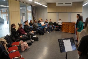 Eine Gruppe von etwa 15 Personen sitzt in einem Raum mit Holzwänden und großen Fenstern. Sie sind auf Stühlen in einem Halbkreis angeordnet und schauen auf zwei Personen, die vor einem Klavier stehen und sprechen. Auf einem Notenständer vor ihnen liegt ein Blatt mit Noten. Der Raum wirkt hell und modern.