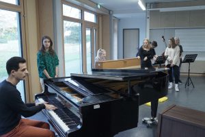 Eine Gruppe von fünf Personen in einem Probenraum. Ein Mann spielt Klavier, während eine Frau in einem grünen Oberteil singt. Im Hintergrund stehen drei weitere Frauen, die offenbar mit dem Gesang üben. Ein weiteres Klavier ist sichtbar. Der Raum hat große Fenster, die viel Tageslicht hereinlassen.