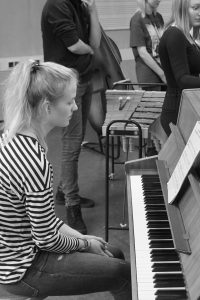 Eine junge Frau mit langen, blonden Haaren sitzt an einem Klavier und schaut nachdenklich nach unten. Sie trägt ein gestreiftes Oberteil und bluejeans. Im Hintergrund sind weitere Personen zu sehen, die an einer musikalischen Aktivität teilnehmen. Der Raum wirkt kreativ und musikalisch.