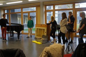 Sechs Personen in einem Raum mit großen Fenstern. Eine Person steht an einem Klavier, während eine andere an einem Podium steht. Die übrigen Personen stehen in einer Gruppe und beobachten. Auf dem Boden liegt eine Kiste. Der Raum hat einen modernen, hellen Charakter.