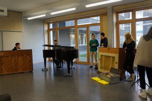 Zwei Klaviere stehen im Vordergrund eines hellen Raumes mit großen Fenstern. Links sitzt eine Person am Klavier, während mehrere Personen im Hintergrund stehen und zuschauen. Eine Person trägt ein grünes Oberteil, die anderen sind in verschiedenen Kleidungsstücken gekleidet. Der Raum hat einen modernen, offenen Charakter.