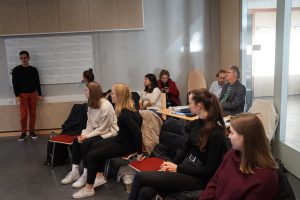 Eine Gruppe von Menschen sitzt in einem Raum mit einem Präsentationsbereich. Ein Mann steht vorne und spricht, während die Zuhörer auf Stühlen sitzen. Die Anwesenden sind unterschiedlich gekleidet, einige tragen helle und andere dunkle Kleidung. Im Hintergrund sind Wände aus Holz und Glas zu sehen.