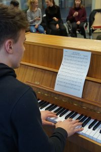 Ein junger Mann sitzt an einem Klavier und spielt, während er auf ein Notenblatt schaut. Im Hintergrund sind einige Personen zu sehen, die ihm zuhören.