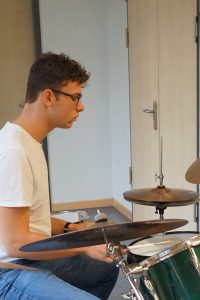 Junger Mann mit Brille, der in einem hellen Raum auf einem Schlagzeug sitzt und spielt. Er trägt ein weißes T-Shirt und Jeans. Im Hintergrund sind Türen und Wände in neutralen Farben sichtbar.