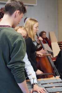 Eine Gruppe von Personen, die an einem musikalischen Workshop teilnehmen. Im Vordergrund sind Jugendliche zu sehen, die auf einem Xylophon spielen. Im Hintergrund sind weitere Teilnehmer, darunter eine Frau mit einem Cello. Die Szene vermittelt eine kreative und gemeinsame Musikatmosphäre.