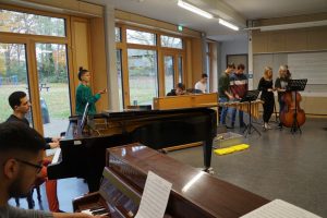 Eine Gruppe von Musikern probt in einem Raum mit großen Fenstern. Im Vordergrund spielt eine Person Klavier, während im Hintergrund mehrere Musiker mit verschiedenen Instrumenten, darunter ein Cello, stehen. Eine weitere Person dirigiert die Probe. Die Atmosphäre ist kreativ und konzentriert.