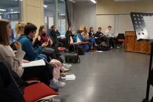 Eine Gruppe von Menschen sitzt in einem Raum auf Stühlen und hört aufmerksam zu. Einige haben Notizblätter oder Notenblätter in der Hand. Im Hintergrund steht ein Klavier, und ein Notenständer ist im Vordergrund sichtbar. Der Raum hat große Fenster und eine helle, freundliche Atmosphäre.