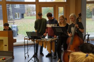 Eine Gruppe von vier Jugendlichen spielt zusammen Musik in einem Raum. Ein Junge sitzt an einem Klavier, während die anderen drei an Notenständern stehen und verschiedene Instrumente spielen: ein Junge spielt auf einem Xylophon, ein Mädchen hat einen Kontrabass, und ein weiterer Junge steht zwischen ihnen. Im Hintergrund sind große Fenster, durch die man einen herbstlichen Garten sehen kann.