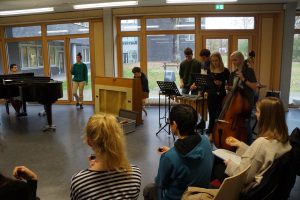 Eine Gruppe von Schülern spielt gemeinsam Musik in einem Raum mit großen Fenstern. Im Vordergrund sitzen einige Schüler, während im Hintergrund Musiker an einem Klavier, einem E-Piano und anderen Instrumenten stehen. Die Atmosphäre ist kreativ und gemeinschaftlich.