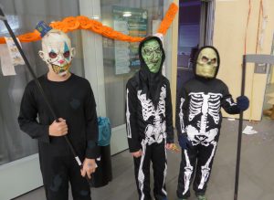 Drei Kinder in Halloween-Kostümen stehen vor einer Tür. Das erste Kind trägt eine Maske mit einem gruseligem Gesicht und ein schwarzes Outfit. Das zweite Kind hat eine grüne Maske und ein schwarzes Kostüm mit einem Skelettmuster. Das dritte Kind trägt eine Schädelmaske und ebenfalls ein schwarzes Skelettkostüm. Im Hintergrund sind orangefarbene Dekorationen zu sehen.