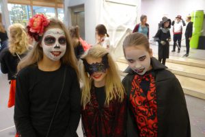 Drei Kinder in Halloween-Kostümen stehen zusammen und lächeln. Sie haben gruselige Schminke im Gesicht: zwei haben weiße Gesichter mit dunklen Augen und Lippen, während das dritte Kind eine schwarze Maske trägt. Die Kinder tragen verschiedene schwarze und rote Kostüme. Im Hintergrund sind weitere Personen in Kostümen zu sehen.