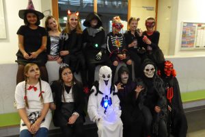 Eine Gruppe von Kindern und Jugendlichen in verschiedenen Halloween-Kostümen. Sie sitzen und stehen zusammen in einem Raum mit hellen Wänden. Die Kostüme umfassen Hexen, Skelette, Geister und andere gruselige Figuren. Einige tragen Masken und Schminke, die ihre Gesichter verzieren. Die Stimmung ist festlich und verspielt.