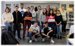 Gruppe von 17 Personen in einem Raum, die lächelnd zusammenstehen. Einige halten Geschenke oder Kuchen in den Händen. Im Hintergrund sind Regale und Stühle sichtbar. Die Personen sind unterschiedlich gekleidet und scheinen sich in einer freundlichen Atmosphäre zu befinden.
