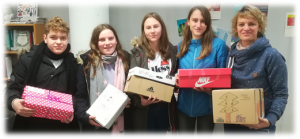 Fünf Personen stehen in einer Innenräume, mit unterschiedlichen Geschenken in den Händen. Ein Junge links hält eine gepunktete Geschenkbox, während die anderen drei Mädchen in der Mitte jeweils eine Geschenkbox halten. Eine Frau rechts trägt eine rote Geschenkbox. Alle lächeln und wirken freundlich.