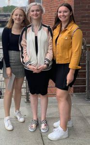 Drei junge Frauen stehen lächelnd nebeneinander auf einer Straße. Die erste trägt ein kariertes Kleid und weiße Sneaker, die zweite hat eine hellrosa Jacke und schwarze Kleidung an, und die dritte trägt eine gelbe Jacke und einen schwarzen Rock. Im Hintergrund sind Ziegelwände und ein Gehweg sichtbar.