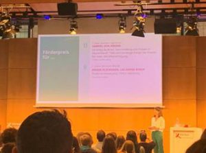 Eine Präsentation auf einer Bühne, mit einem Text auf einem großen Bildschirm im Hintergrund. Im Vordergrund steht eine Person, die spricht. Der Raum ist mit Publikum gefüllt, das in Richtung der Präsentation blickt. Die Bildschirmtexte sind teilweise sichtbar und scheinen sich mit einem Thema über Förderung zu befassen. Die Atmosphäre ist informativ und engagiert.