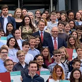 Gruppe von Schülern und Lehrern, die auf einer Treppe stehen und stolz ihre Zertifikate oder Urkunden halten. Die Szene zeigt eine Vielzahl von Gesichtern und Kleidungsstilen, die die Diversität der Gruppe unterstreichen. Im Hintergrund sind moderne Gebäude zu sehen.