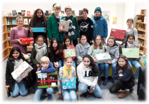 Gruppe von Kindern und Jugendlichen, die in einer Bibliothek stehen und sitzen. Sie halten bunt verpackte Geschenke in den Händen. Im Hintergrund sind Bücherregale sichtbar. Einige Personen tragen winterliche Kleidung.