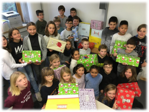 Eine Gruppe von Kindern und Jugendlichen steht in einem Raum und hält bunte, liebevoll verpackte Geschenke in den Händen. Sie lächeln und posieren für das Foto, während einige Geschenke auf dem Boden und in der Umgebung sichtbar sind. Im Hintergrund sind weitere Geschenke und ein Drucker zu sehen.