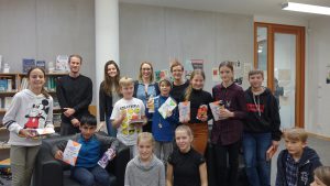 Eine Gruppe von Kindern und Erwachsenen steht in einer Bibliothek. Die Kinder halten verschiedene Bücher und Materialien in den Händen. Im Hintergrund sind Regale mit Büchern sichtbar. Die Atmosphäre wirkt freundlich und einladend.
