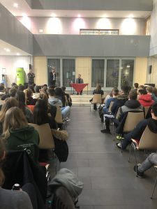 Eine Gruppe von Menschen sitzt in einem modernen Raum auf Stühlen, während zwei Personen auf einer Bühne an einem Tisch sprechen. Im Hintergrund sind Fenster zu sehen, die Tageslicht hereinlassen. Die Anwesenden sind aufmerksam und schauen in Richtung der Bühne.