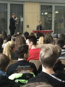 Zwei Männer stehen und sitzen auf einer Bühne während einer Veranstaltung. Der Mann im Anzug steht und spricht zu einem Publikum, das aus jungen Menschen besteht, die in einem großen Raum sitzen. Der Mann, der sitzt, hat einen Tisch vor sich. Im Hintergrund sind Fenster und eine festliche Dekoration zu sehen.