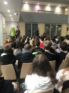 Eine Gruppe von Schülern sitzt in einem Raum und hört einem Vortrag zu. Im Vordergrund stehen zwei Personen, die sprechen, während die Zuhörer aufmerksam schauen. Der Raum hat große Fenster und ist hell erleuchtet.