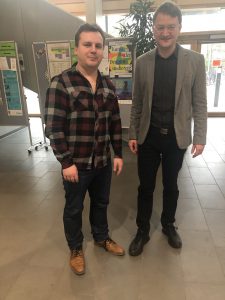 Zwei Männer stehen in einem hellen Innenraum, umgeben von Informationsplakaten. Der links stehende Mann trägt ein kariertes Hemd und Jeans, während der rechts stehende Mann einen grauen Blazer über einem schwarzen Hemd trägt. Beide schauen freundlich in die Kamera.