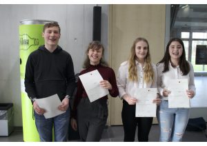 Vier Jugendliche stehen nebeneinander und halten jeweils ein Dokument in der Hand. Sie lächeln und scheinen stolz zu sein. Im Hintergrund ist eine grüne Säule und eine Wand sichtbar. Die Gruppe befindet sich anscheinend in einem Innenraum.