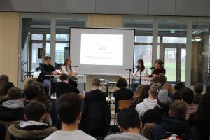 Eine Gruppe von vier Personen sitzt an einem Tisch auf einer Bühne, während sie vor einem Publikum sprechen. Im Hintergrund ist ein Bildschirm sichtbar, auf dem eine Präsentation angezeigt wird. Das Publikum, bestehend aus Jugendlichen, sitzt in Reihen und beobachtet die Diskussion. Die Umgebung ist hell und modern gestaltet, mit großen Fenstern.