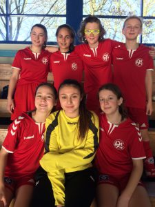 Eine Gruppe von sieben Mädchen in roten Fußballtrikots steht und sitzt zusammen in einer Sporthalle. Einige Mädchen tragen Sonnenbrillen. Die Atmosphäre wirkt freundlich und sportlich. Im Hintergrund sind Fenster, durch die Tageslicht strömt.